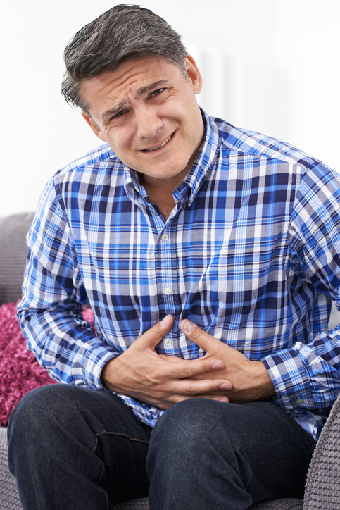 Dor abdominal, cólicas, inchaço, movimentos intestinais ruidosos no abdómen: Getty Images SII - homem com dores abdominais, cólicas, segurando o abdómen, camisa de xadrez azul, sentado no sofá