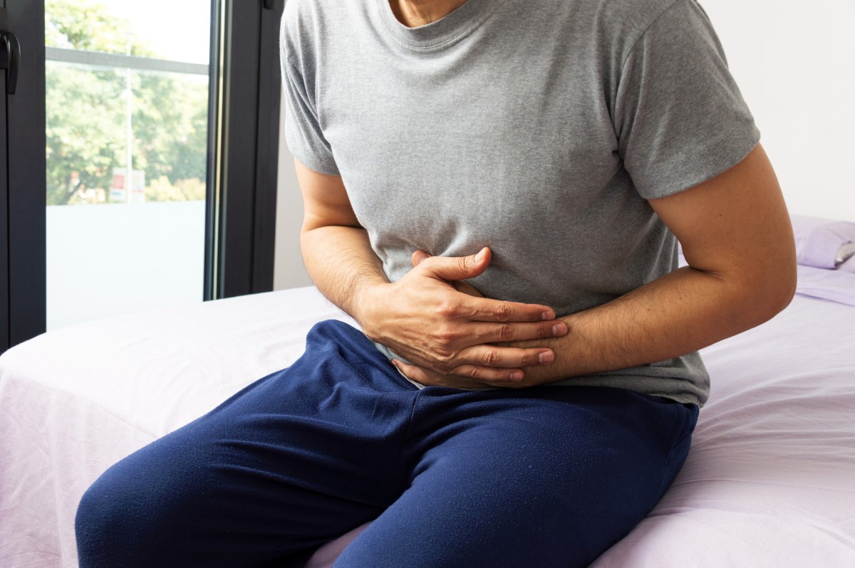O estômago de um homem dói-lhe, está a segurá-lo com as mãos e está sentado na cama de pijama.