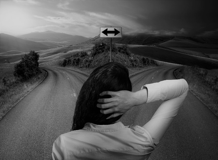 Escolher a direção certa é a coisa mais importante. Fonte: thinkstock photos mulher de pé na encruzilhada, de costas, segurando a parte de trás da cabeça com a mão, há uma seta na encruzilhada em ambas as direcções