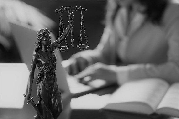 Uma carga de trabalho elevada leva ao stress Fonte: thinkstock photos uma mulher sentada a uma mesa com um livro à sua frente e uma estátua da justiça em primeiro plano sobre a mesa