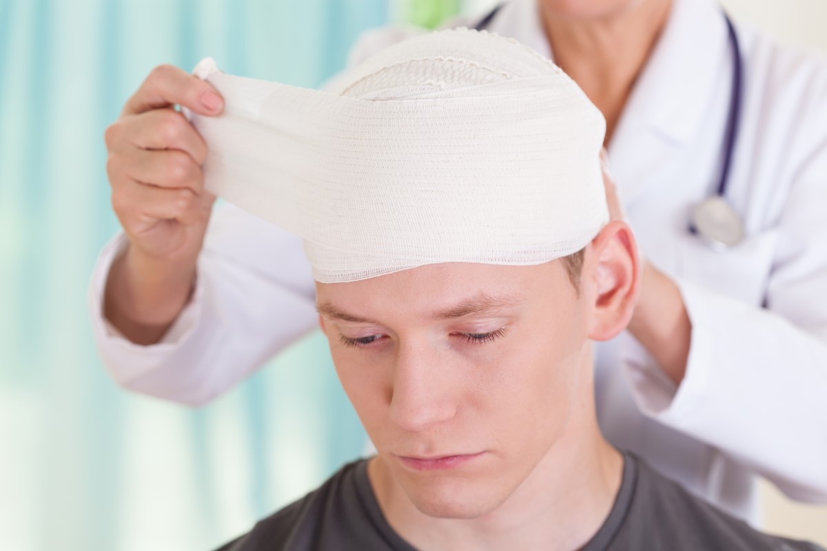 Após um traumatismo craniano, é importante monitorizar o estado e o aparecimento de problemas de saúde: Getty Images Dor de cabeça após acidente, cabeça enfaixada, médico trata rapaz com traumatismo craniano