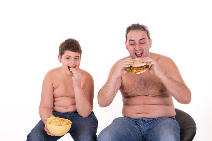 Hábitos alimentares inadequados e obesidade dos pais. Fonte da foto: Getty Images o rapaz e o seu pai comem os alimentos errados, comem hambúrgueres, são obesos