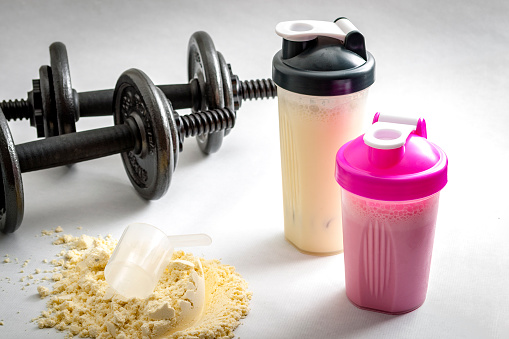 Os suplementos alimentares existem em vários sabores. Fonte da fotografia: Getty Images Dois halteres para uma mão, colocados ao lado das bebidas proteicas, em duas taças, e uma proteína em pó com uma colher de medida ao lado.