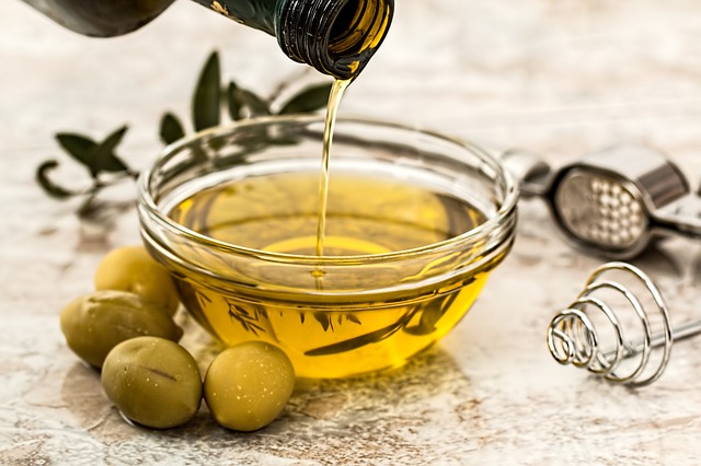 O azeite é um componente importante da dieta mediterrânica. Fonte: Pixabay Deita-se azeite de uma garrafa de vidro para uma taça de vidro e colocam-se azeitonas verdes à volta da taça.