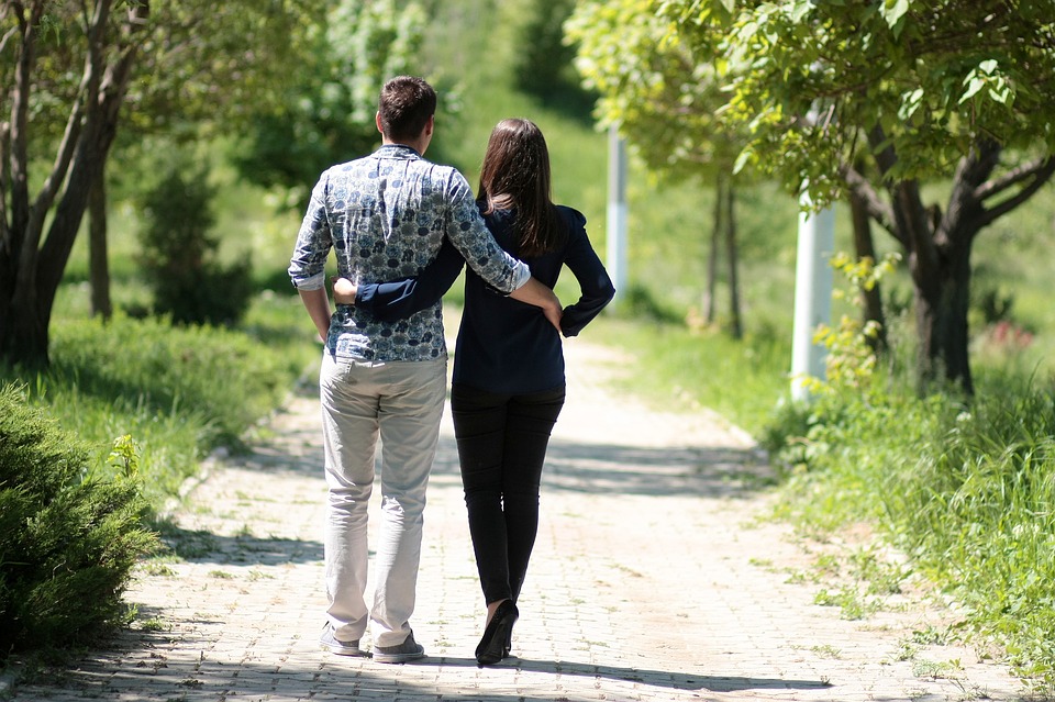 Por vezes, até um simples passeio pode aliviar o stress melhor do que qualquer outra coisa. Fonte: Thinkstock Casal a caminhar num trilho natural, abraçados pela cintura, com árvores verdes e relva verde.
