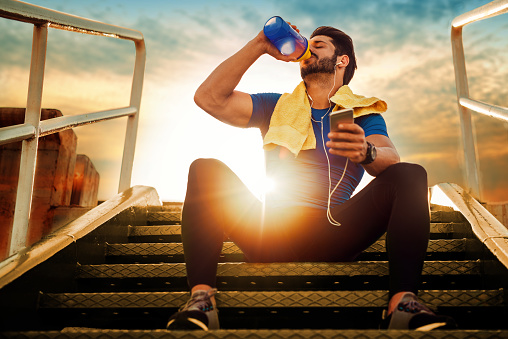 Alguns suplementos alimentares podem ser tomados durante o treino. Fonte da fotografia: Getty Images Um atleta sentado nas escadas. Pôr do sol. Beber um suplemento nutricional de um frasco.