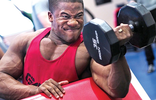 As proteínas são um bom suplemento alimentar para aumentar a massa muscular. Fonte da foto: Pixabay Homem de camisola de alças vermelha, a fazer bíceps com uma barra de um braço.