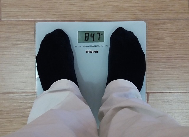 Para calcular o seu IMC, tudo o que precisa de saber é o seu peso e altura. Fonte: Thinkstock Um homem, vestindo calças de cor clara e meias pretas, está em cima de uma balança digital, medindo o seu peso. O mostrador indica 84,7 kg. A balança é branca, de vidro, sobre um chão flutuante de cor clara.