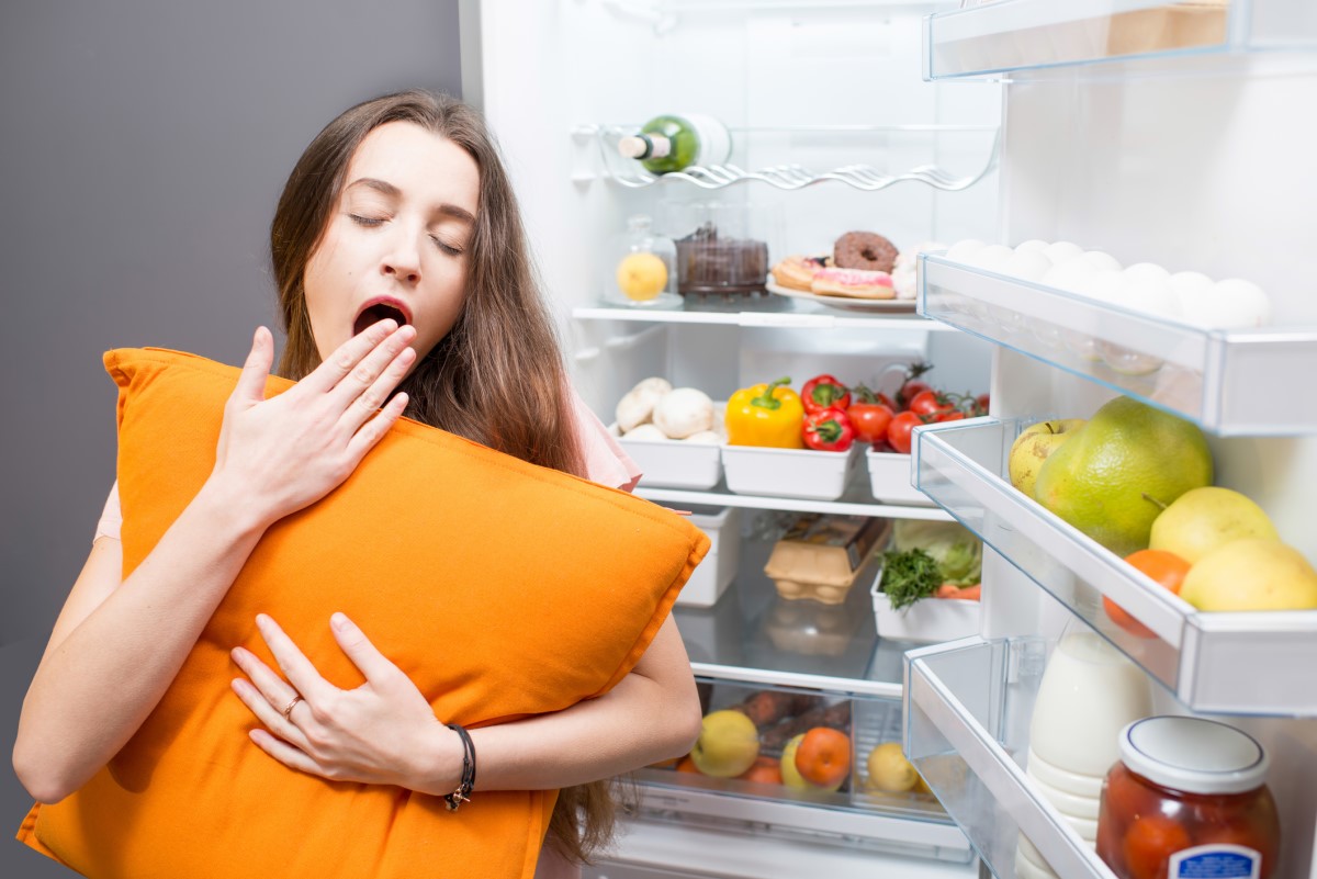 Uma mulher a dormir junto a um frigorífico, ilustrando os hábitos alimentares como um fator que influencia a qualidade do sono.