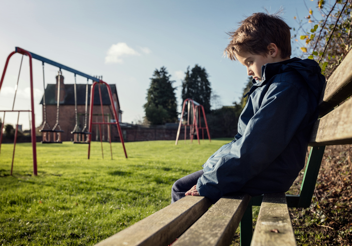 rapaz sentado sozinho num banco, triste, com emoções negativas, parque infantil, relva, baloiço
