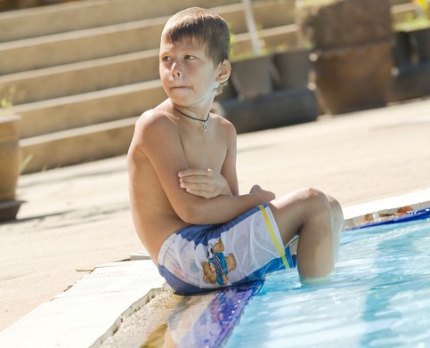 As crianças devem ter cuidado mesmo quando tomam banho no verão. Fonte da fotografia Getty Images O rapaz está sentado junto à piscina, com os pés na água. Tem frio.